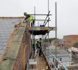Builders standing on a scaffold working on repairing a historic red brick wall and slate roof.