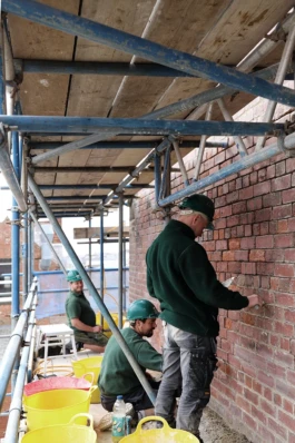 Builders standing on a scaffold working on re-pointing a historic red brick wall.