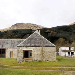 A renovated historic stone barn with a large square window and mountains in the background.