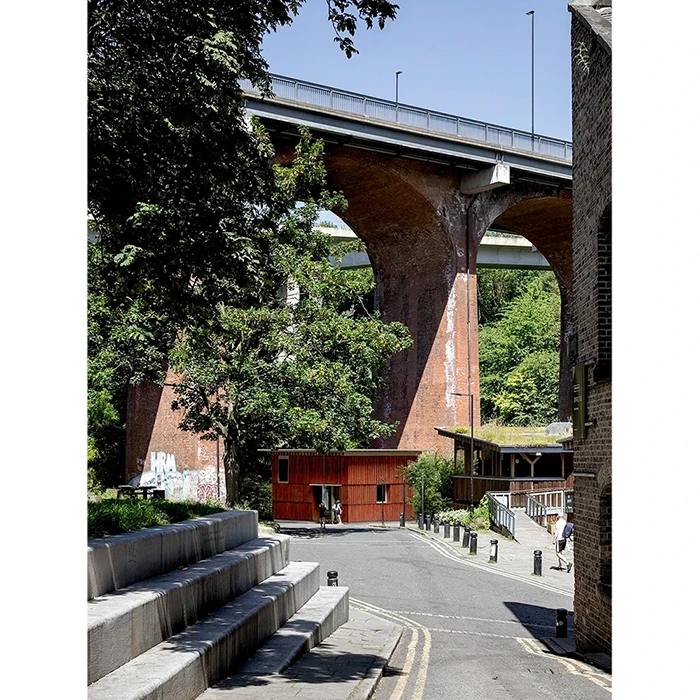 Topo Architects — A red timber building below a large, arched, vehicular bridge.