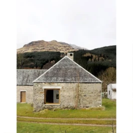 A renovated historic stone barn with a large square window and mountains in the background. 